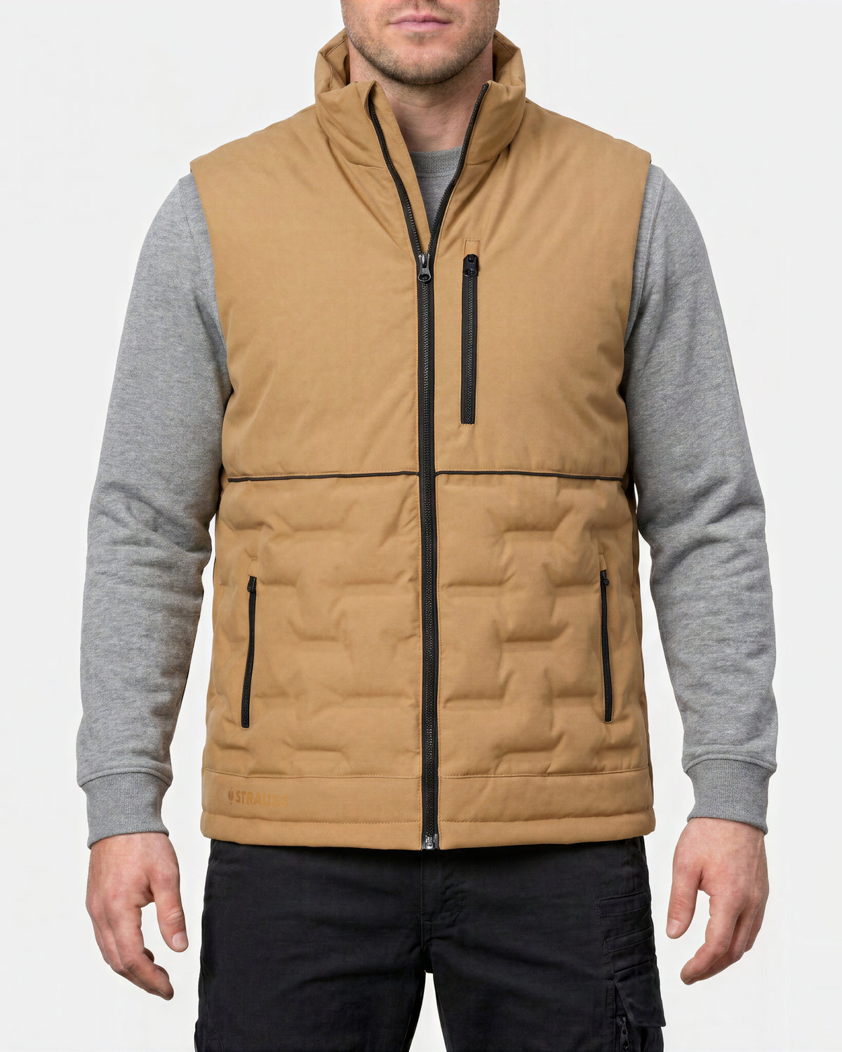A man wearing a tan insulated zip-front vest over a gray long-sleeve shirt and black pants, shown from the lower face to upper thighs against a light gray studio background.