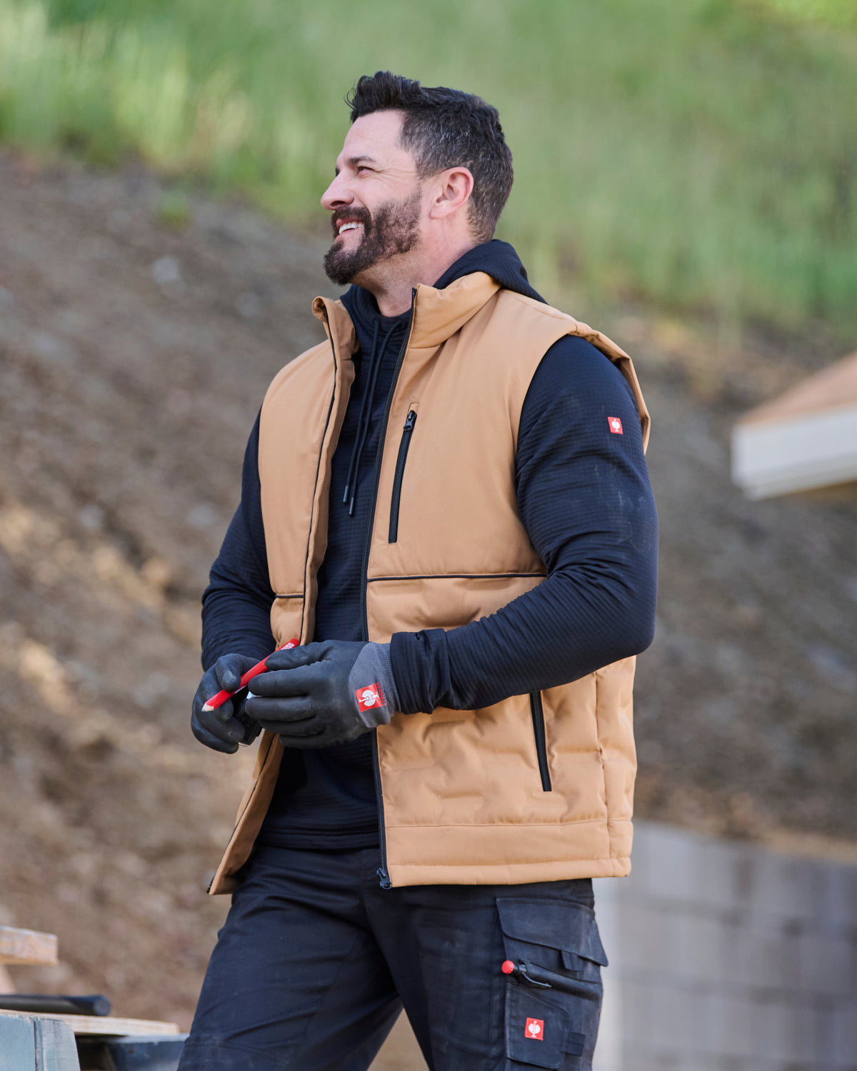 A man wearing a tan insulated work vest over a black hoodie and black work gloves, standing outdoors against a blurred background of dirt and grass.