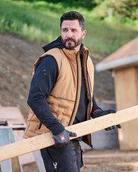 A man wearing a tan quilted puffer vest over a black hoodie and work gloves carries a long wooden board on an outdoor construction site.