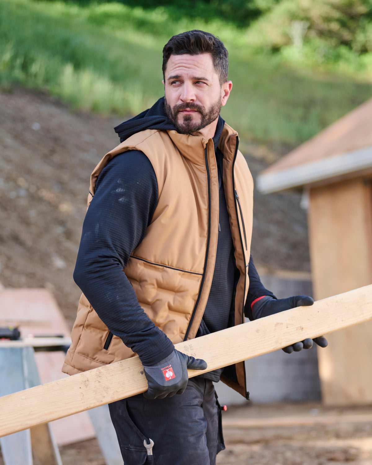 A man wearing a tan quilted puffer vest over a black hoodie and work gloves carries a long wooden board on an outdoor construction site.