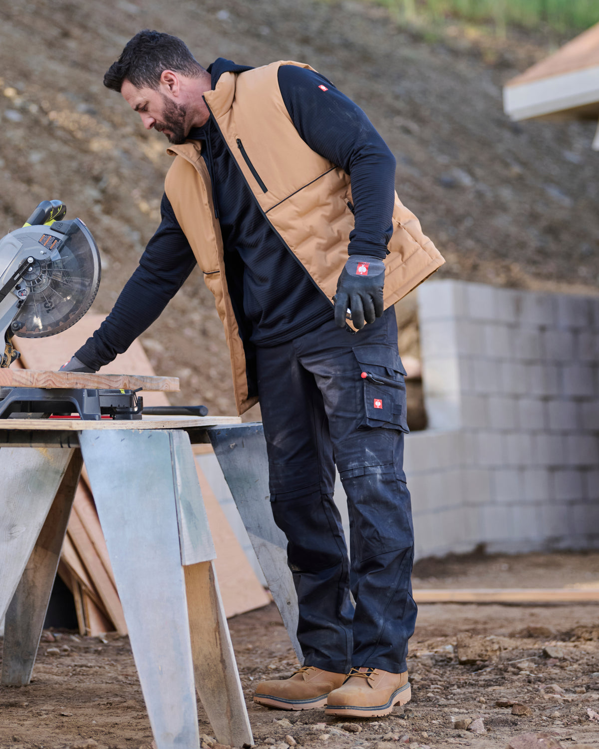 A man wearing a tan work vest over a black long-sleeve shirt and dark cargo pants stands at a workbench with a circular saw in a construction yard setting.