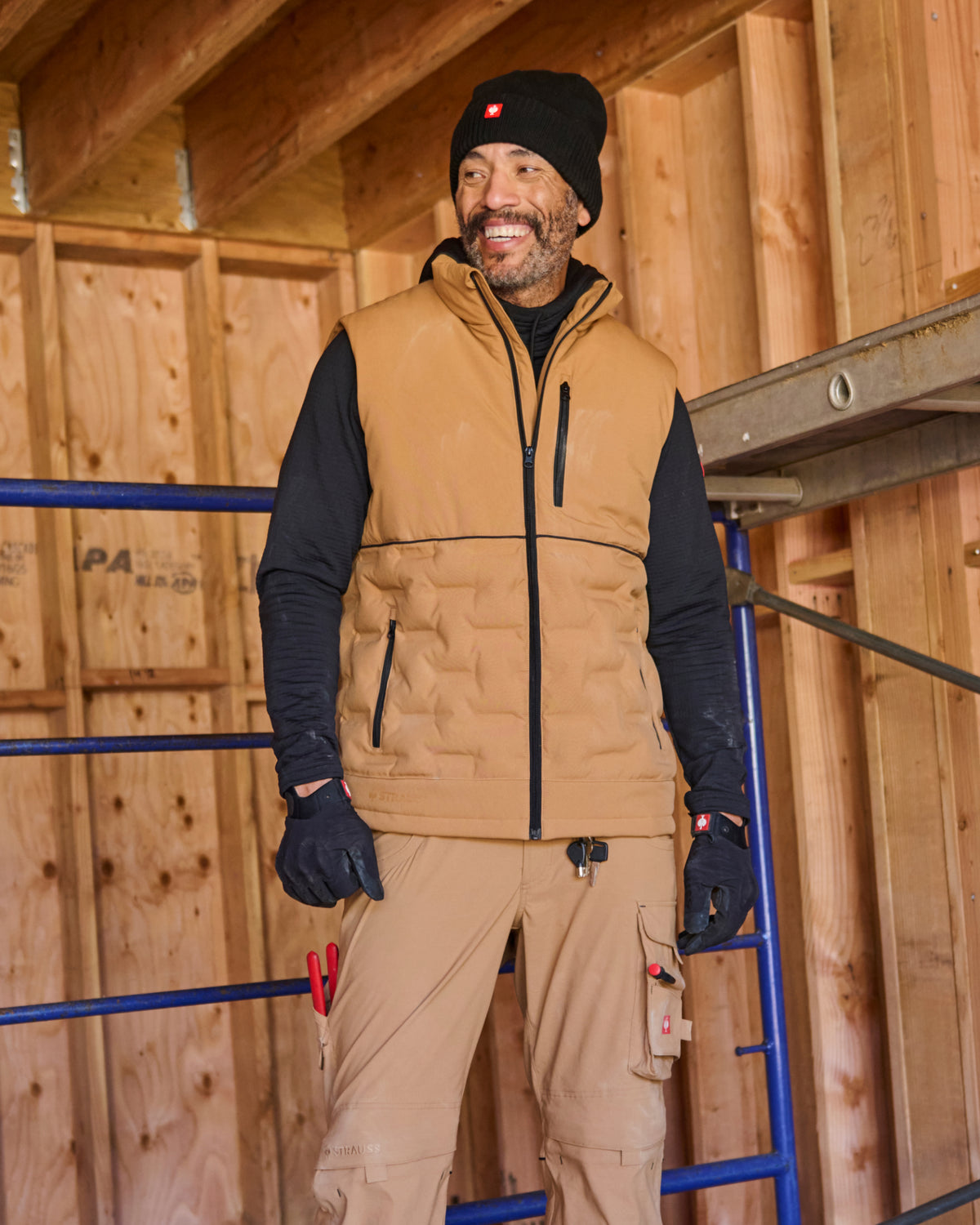 A smiling man wearing a tan insulated work vest over a black long-sleeve shirt, tan work pants, black gloves, and a black beanie stands indoors against a wood-framed wall background.