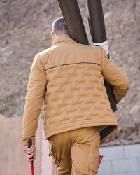 A man viewed from behind wearing a tan quilted puffer jacket and matching tan cargo pants, carrying two large dark poles over his shoulder in an outdoor setting.
