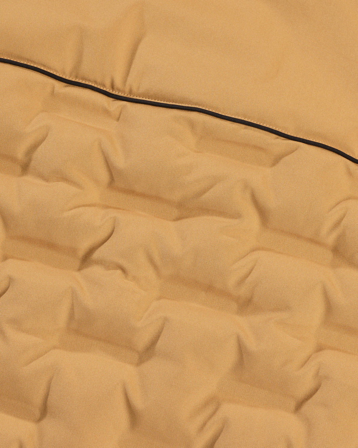 Close-up of a quilted fabric surface in a warm camel tan color with a thin black piping detail running horizontally.