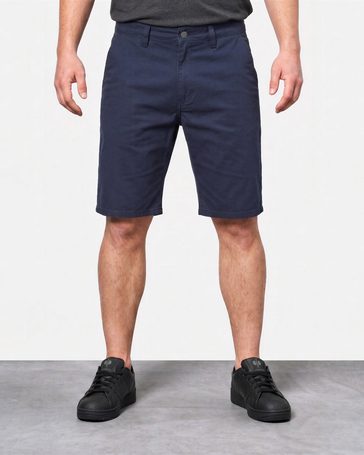 A person wearing navy blue knee-length shorts, a dark gray t-shirt, and black sneakers, standing against a white wall and gray floor.