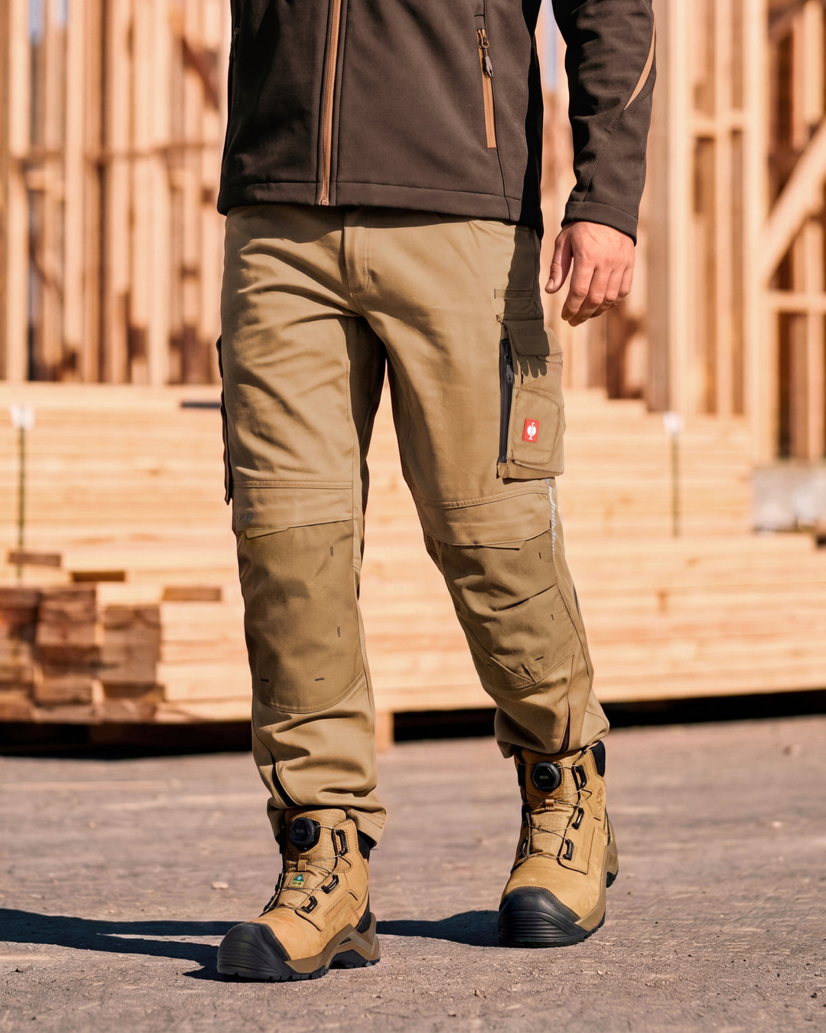 A person wearing tan work pants with cargo pockets and tan work boots, standing outdoors in front of a wooden construction site. The jacket is partially visible.
