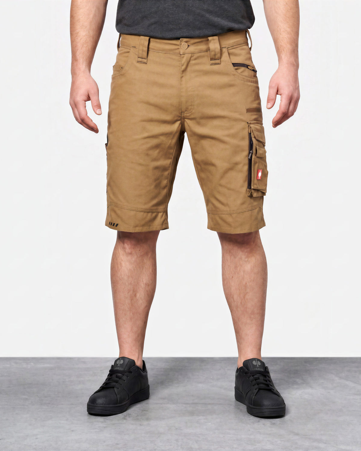 A person wearing tan cargo shorts with black sneakers, shown from the waist down against a plain light gray studio background.