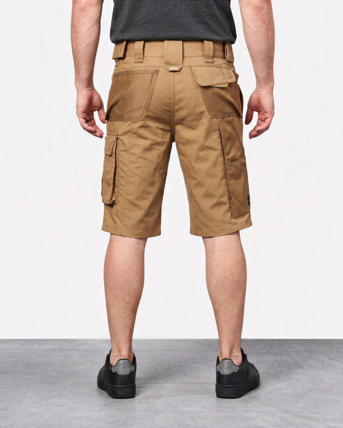 A man viewed from the back wearing tan cargo shorts, a dark gray t-shirt, and black sneakers, standing against a white studio background.