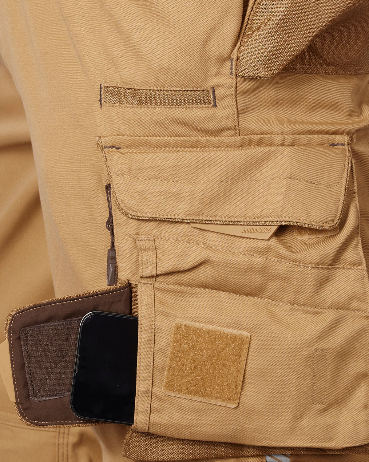 Close-up of a tan work shorts showing a side utility pocket with a brown zippered compartment holding a black smartphone, alongside a tan Velcro patch.
