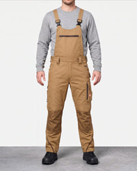 A man wearing tan work bib overalls over a gray long-sleeve shirt, standing against a light gray studio background.