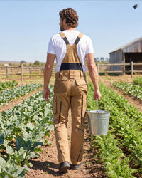 A man viewed from behind walking through a farm field, wearing tan bib overalls over a white T-shirt and carrying a metal bucket.
