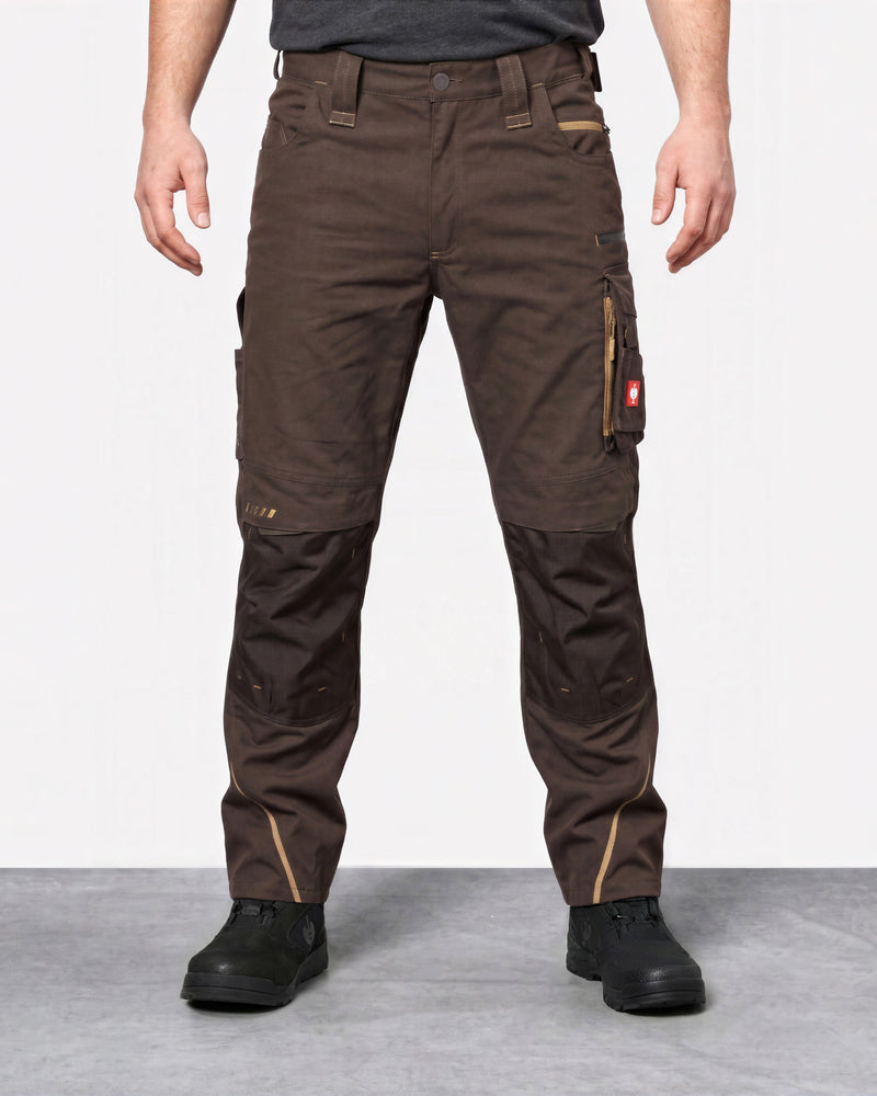 A person wearing dark brown work pants with reinforced knee panels and black work boots, standing against a light gray studio background.