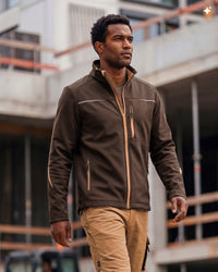 A man wearing a brown zip-up jacket with tan accents and khaki pants, walking in front of a blurred construction site background.
