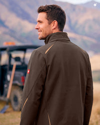 A man wearing a brown athletic jacket with tan contrast stitching, photographed outdoors with a blurred mountain background and a vehicle behind him.