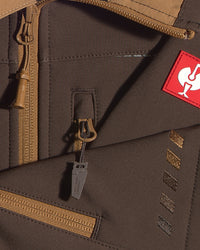 Close-up detail of a brown softshell jacket with tan zippers, a red square patch with a white rooster logo, and a gray zipper pull tag.