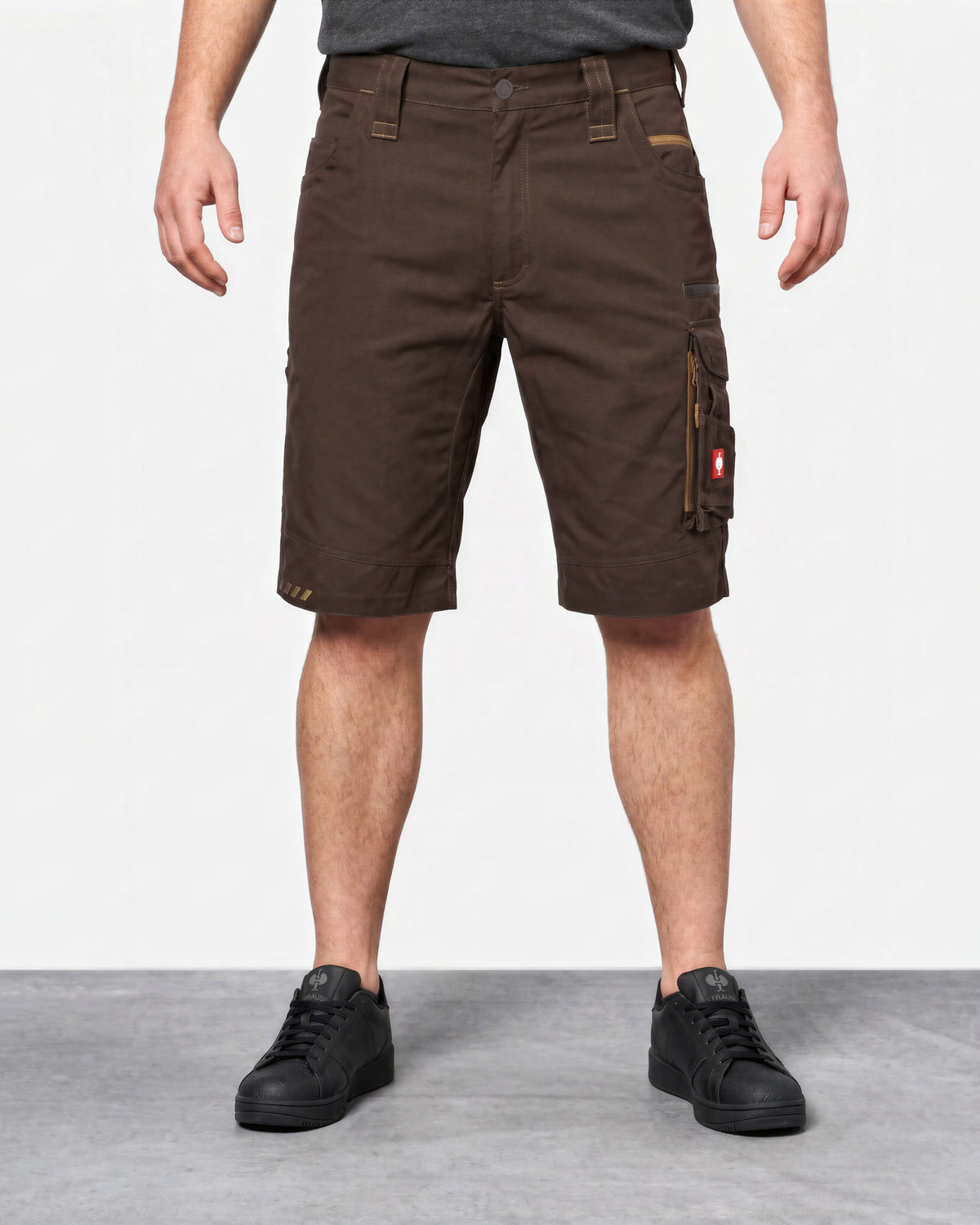 A person wearing dark brown cargo shorts with side utility pockets, paired with black sneakers, shown from the waist down against a light gray studio background.