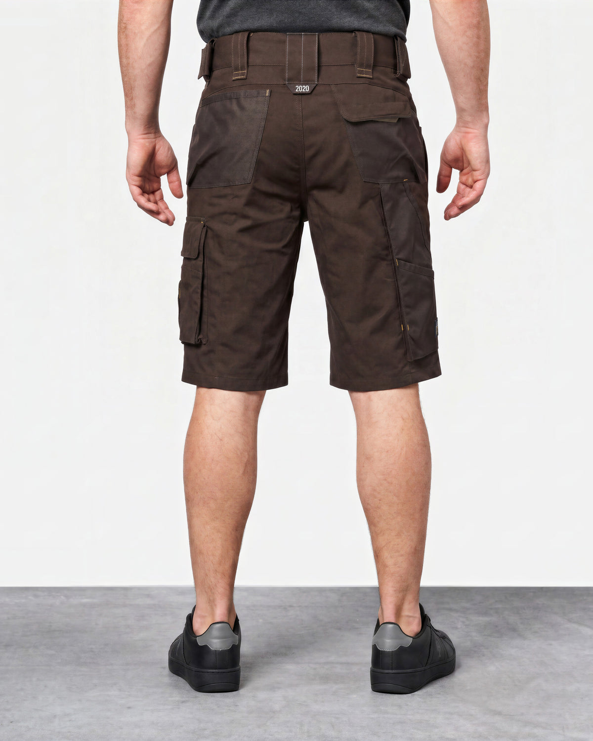 A man viewed from the back wearing dark brown cargo shorts, a dark gray t-shirt, and black sneakers, standing against a light gray studio background.