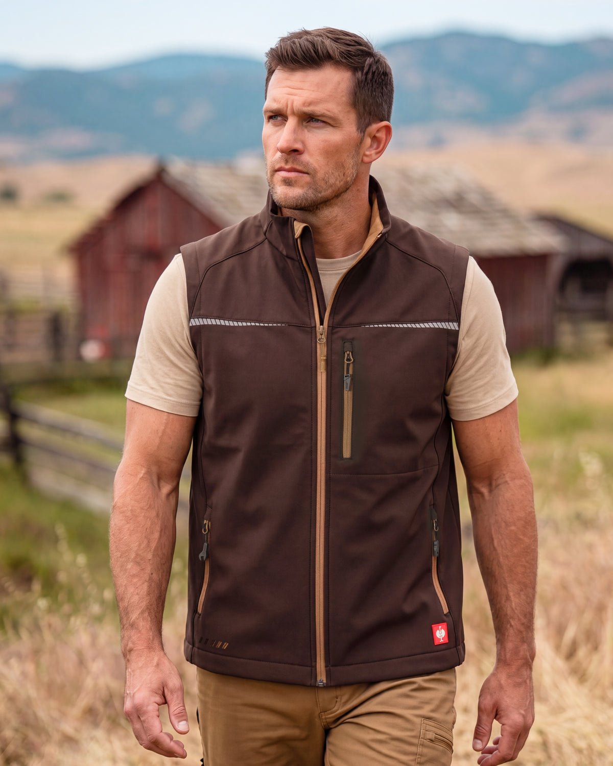 A man wearing a brown insulated zip-front vest over a beige t-shirt and khaki pants, standing outdoors in a rural setting with a red barn and mountains in the background.