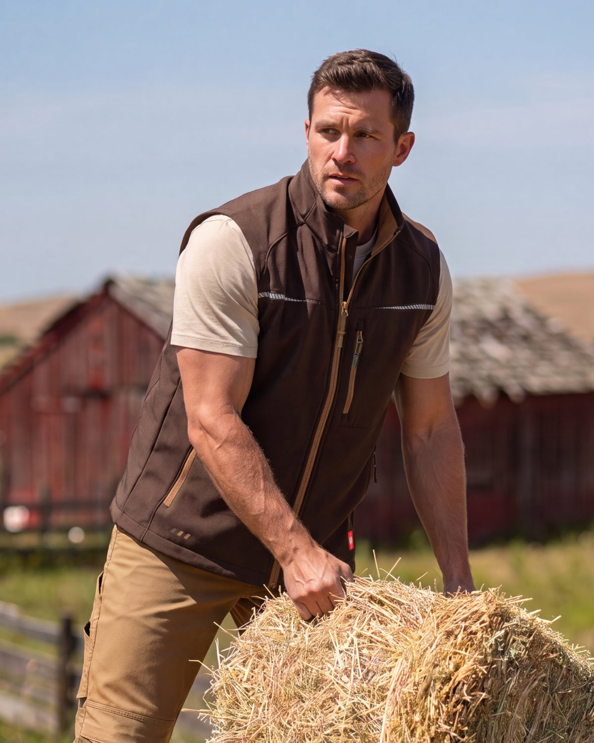 A man wearing a brown zip-front vest over a beige t-shirt and khaki pants, standing outdoors next to a hay bale with red barns in the background.