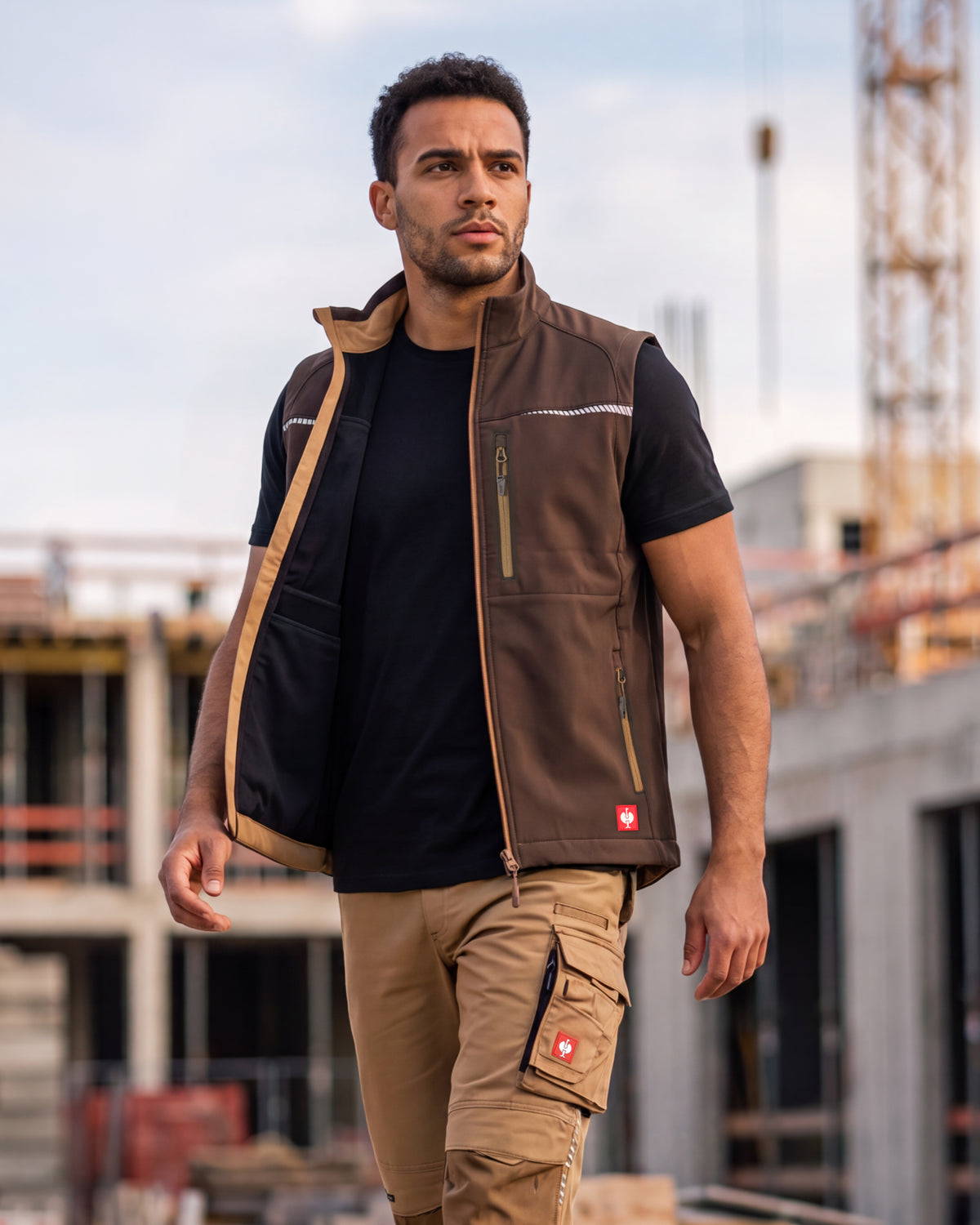 A man wearing a brown work vest over a black t-shirt and khaki cargo pants, standing on a construction site.