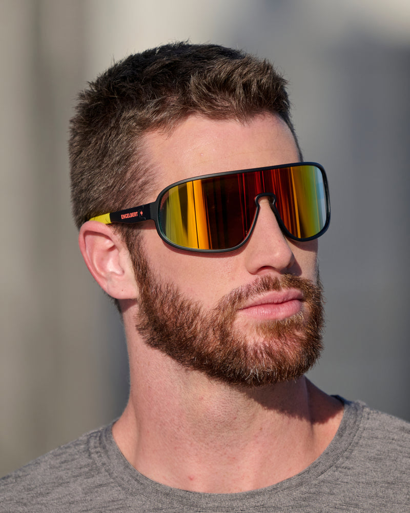A worker wears black and high-vis yellow STRAUSS e.s.ambition race sunglasses with outdoor workwear in bright conditions.
