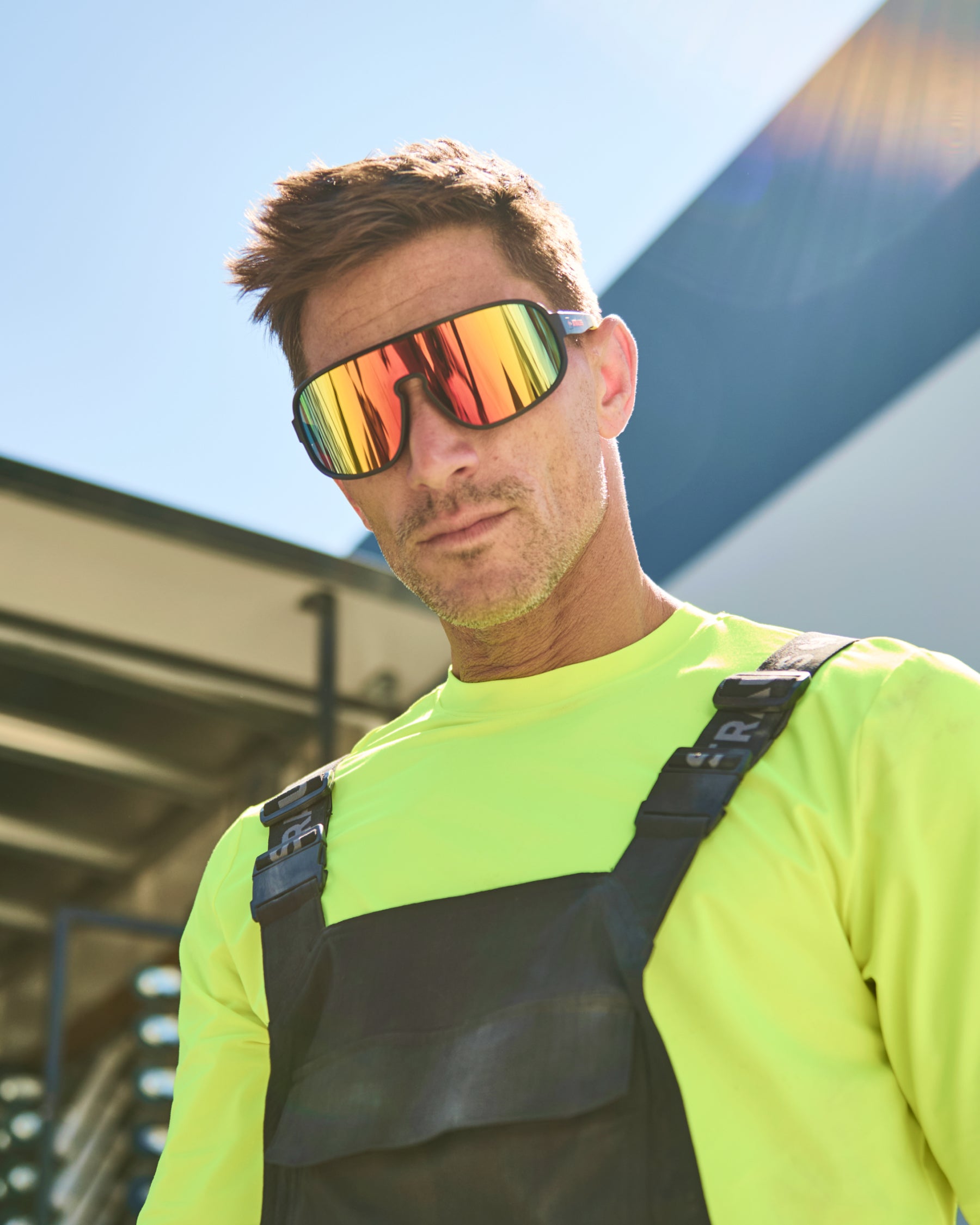 A worker wears black and high-vis yellow STRAUSS e.s.ambition race sunglasses with outdoor workwear in bright conditions.
