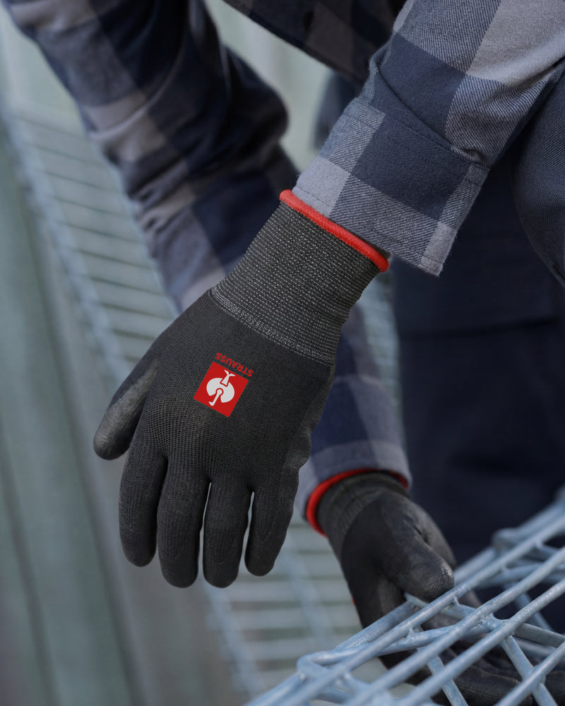 A worker wears black STRAUSS PU micro gloves while gripping a hand tool at a workbench.