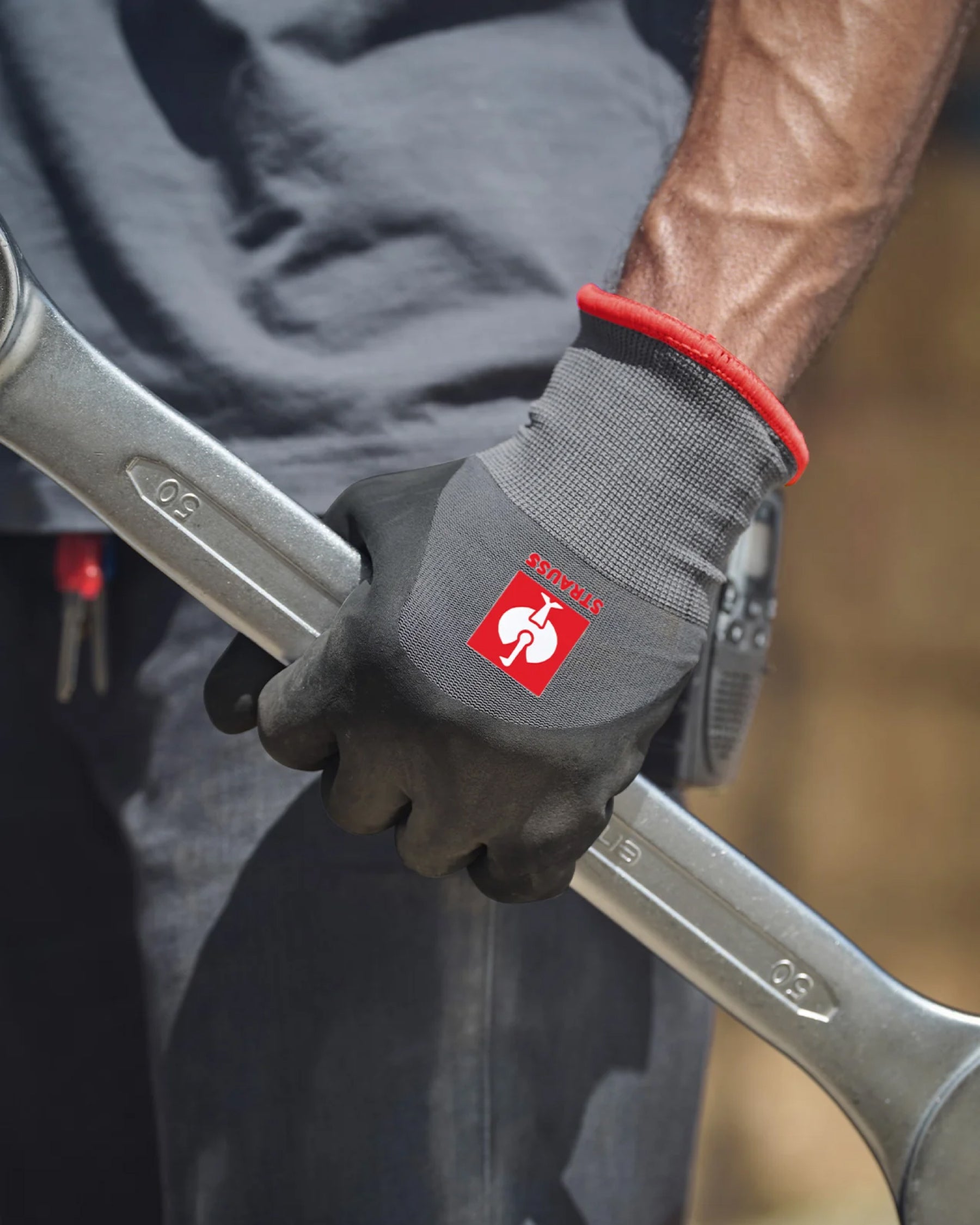 A worker wears black and gray STRAUSS evertouch winter nitrile foam gloves while gripping a hand tool at a workbench.