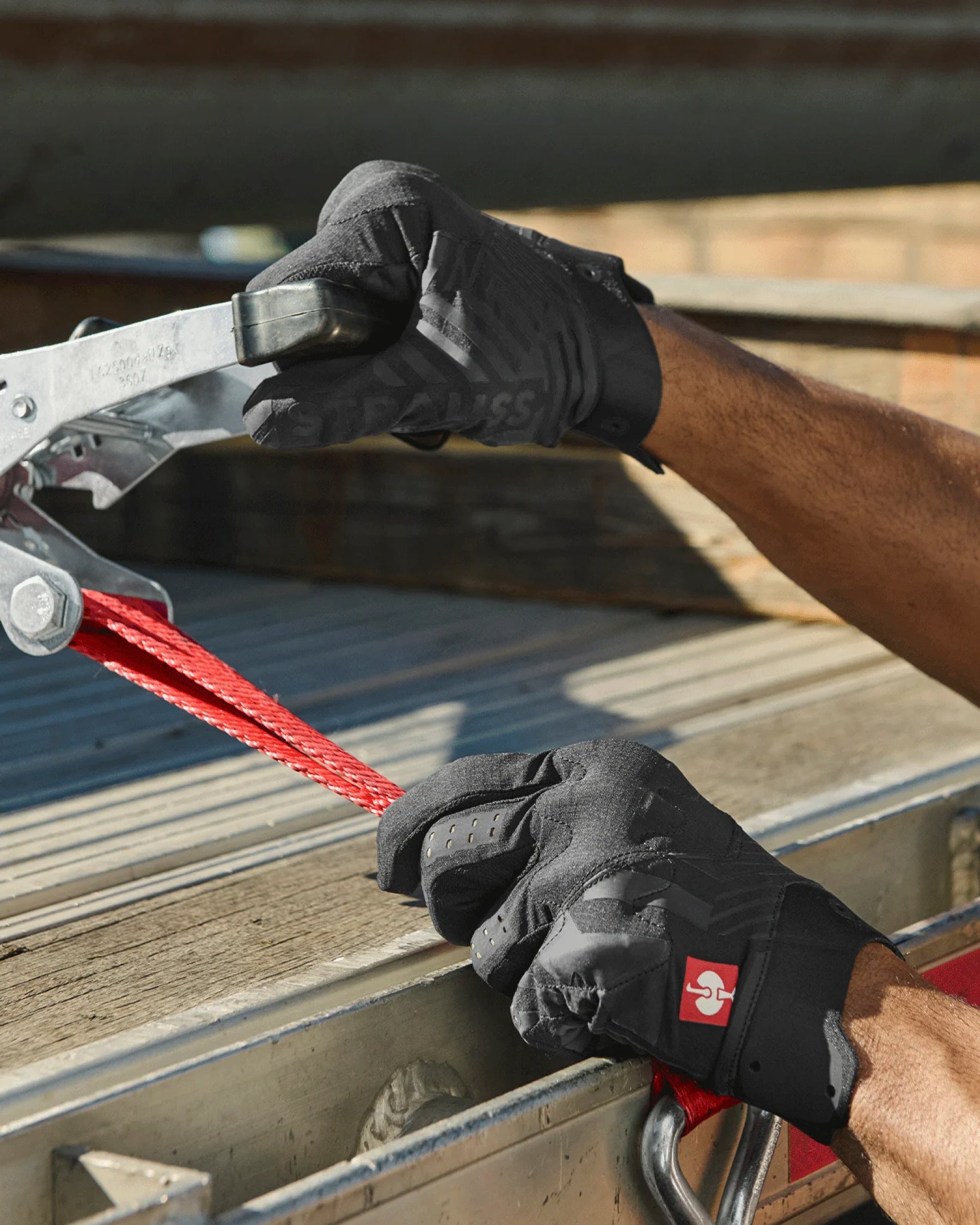 A worker wears black STRAUSS e.s.trail light graphic mechanic gloves while gripping a hand tool at a workbench.