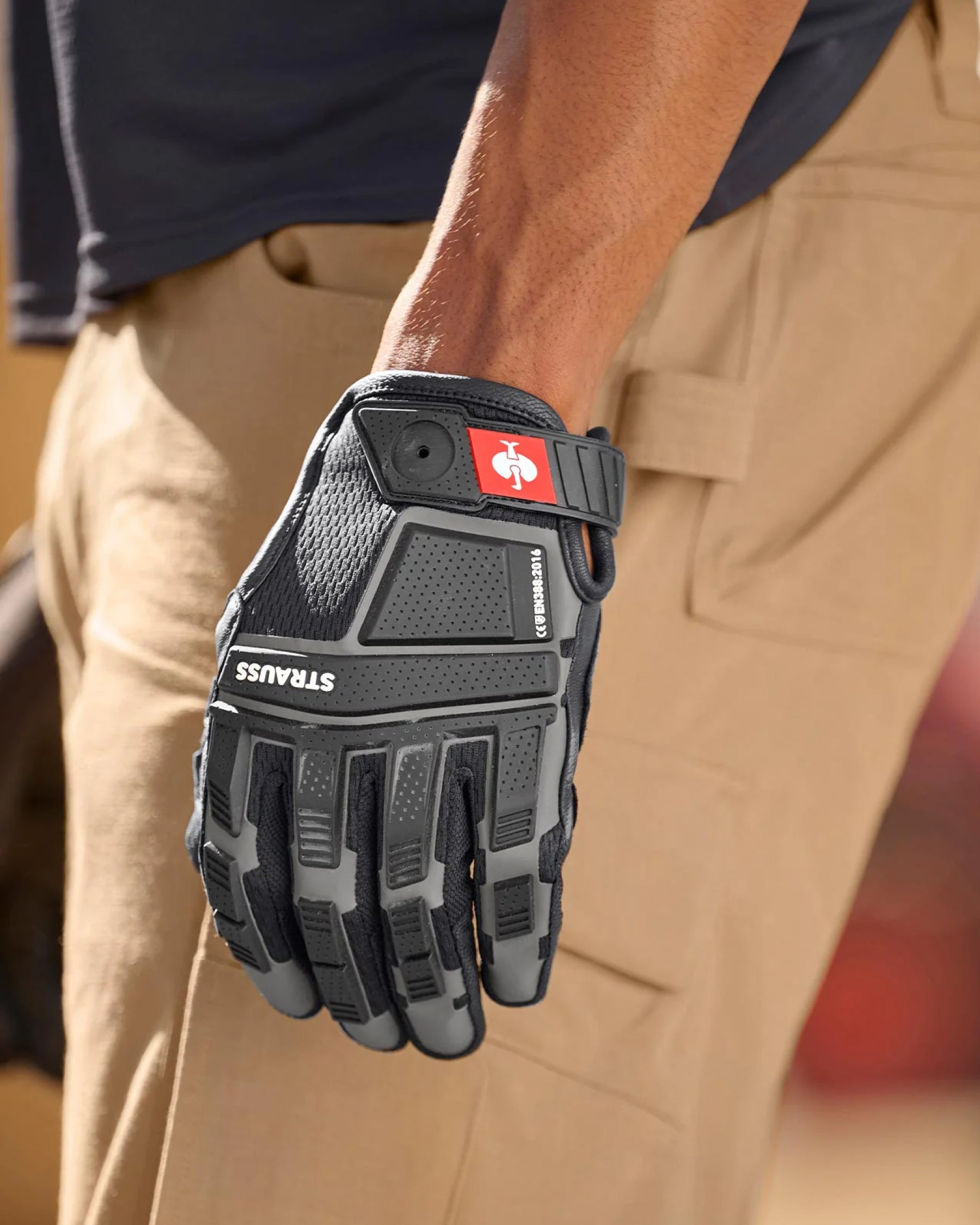 A worker wears black STRAUSS assembly protection gloves while gripping a hand tool at a workbench.