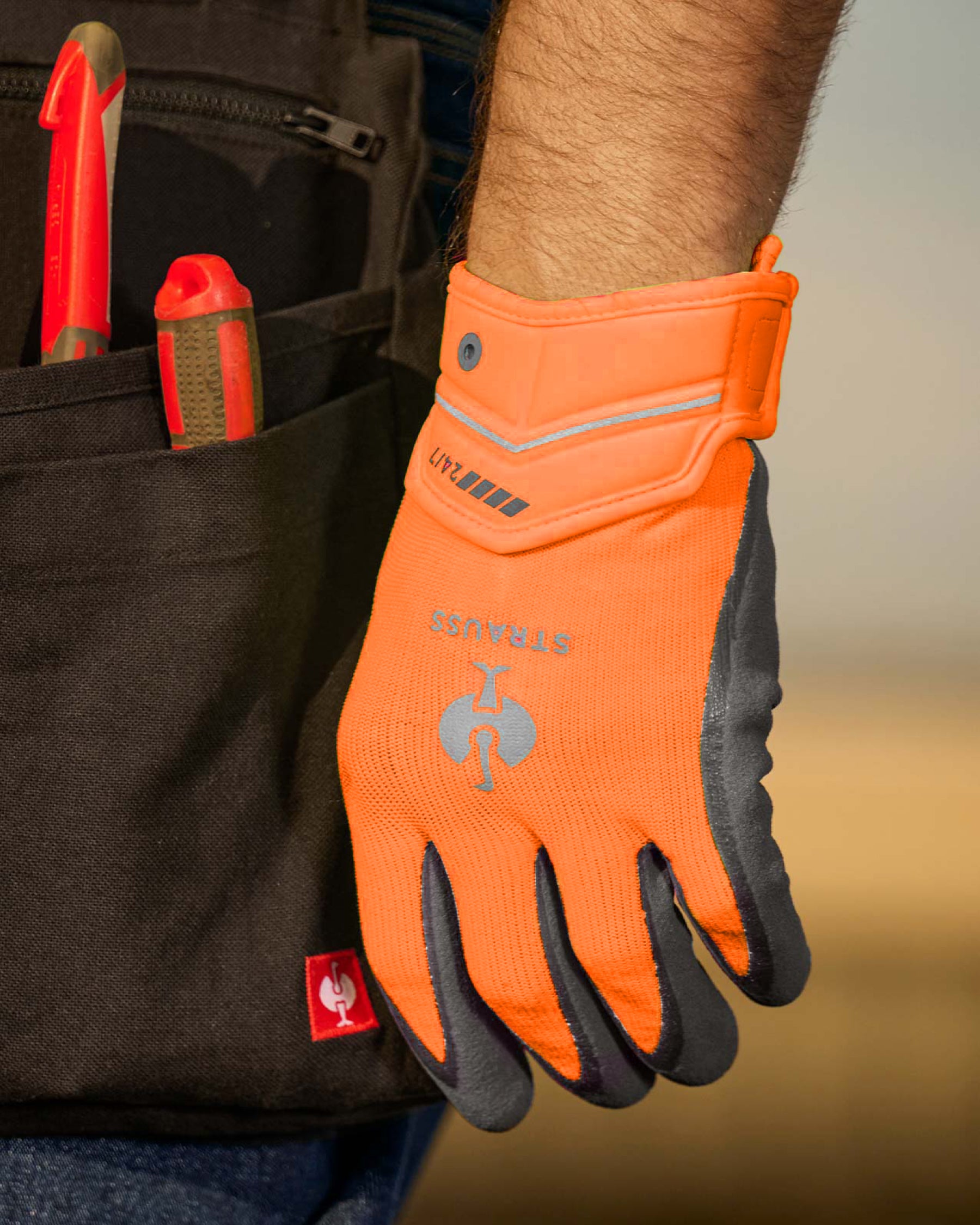 A close work scene shows neon orange and anthracite STRAUSS e.s.motion 24/7 hybrid gloves protecting hands during a task.