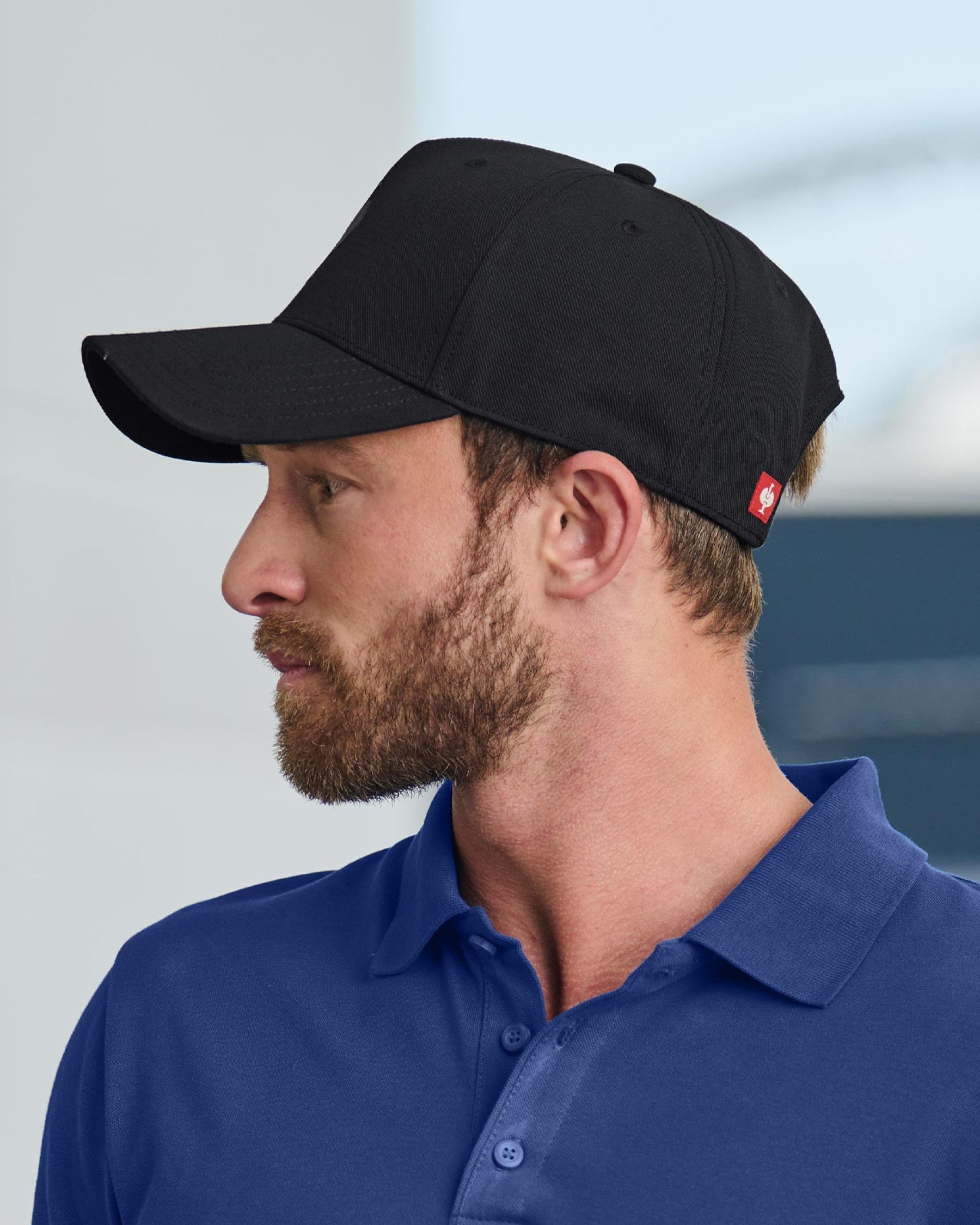 A worker in a black STRAUSS e.s.trail trucker hat stands near jobsite materials in outdoor workwear.
