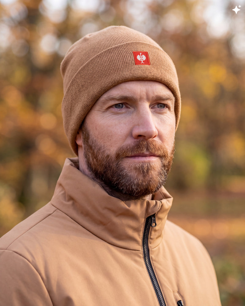 A man wearing a tan knit beanie with a small red square logo patch and a matching tan zip-up jacket, photographed outdoors against a softly blurred autumn background.