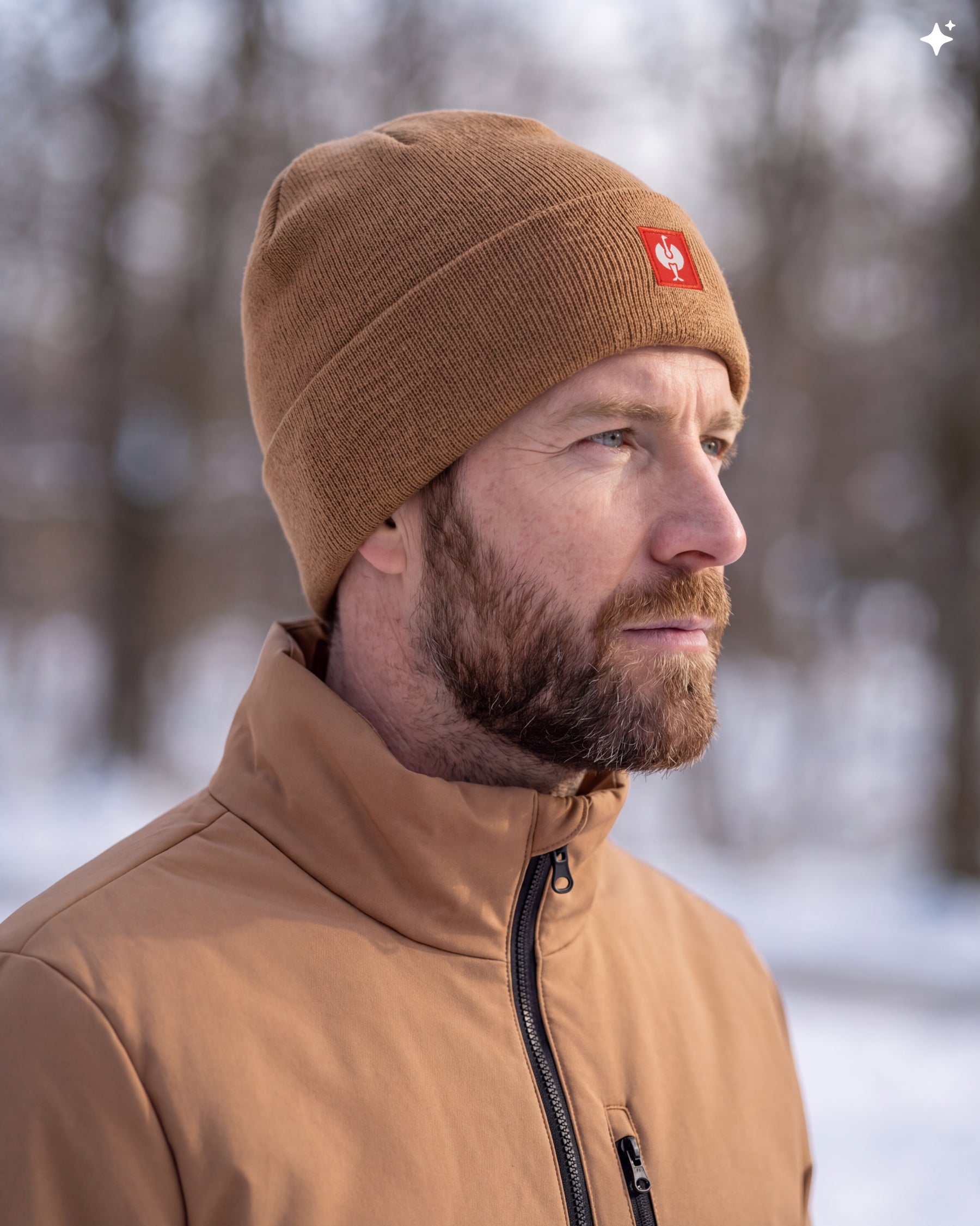A man in a snowy outdoor setting wearing a brown knit beanie with a small red square logo patch and a matching brown jacket.