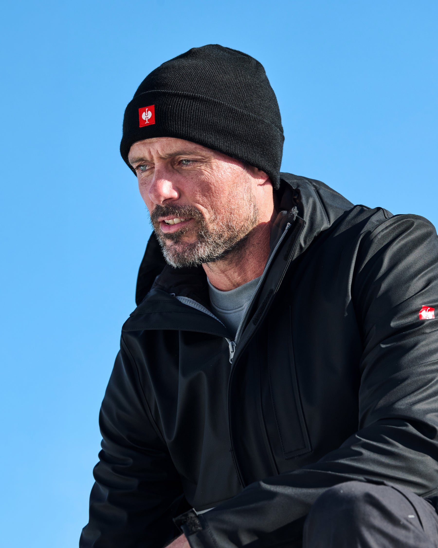 A man wearing a black knit beanie with a red and white logo patch and a black outdoor jacket, photographed against a clear blue sky.