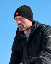 A man wearing a black knit beanie with a red and white logo patch and a black outdoor jacket, photographed against a clear blue sky.