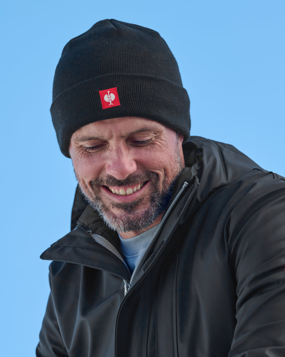 A man wearing a black knit beanie with a small red square logo patch on the front, photographed outdoors against a clear blue sky.