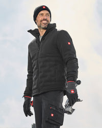 A man wearing a black Swissguard jacket, matching black pants, black gloves, and a black beanie with a red Swiss cross logo, holding a camera against a bright, overcast sky.