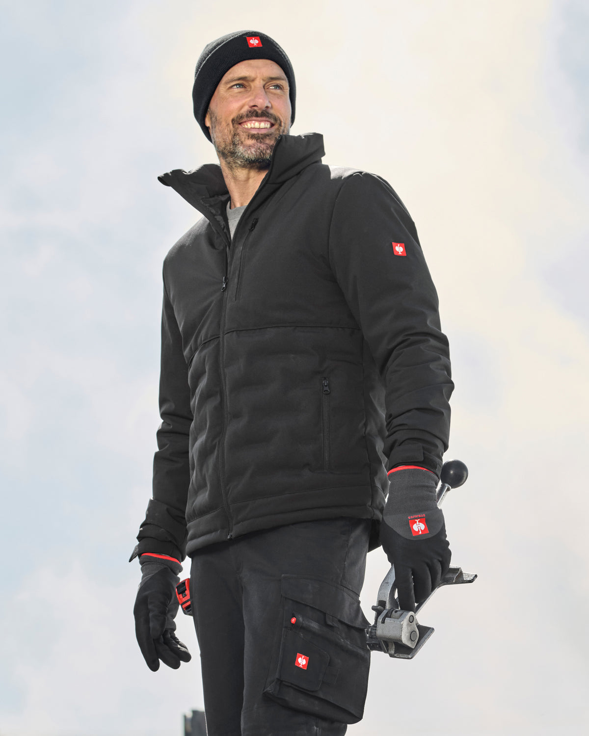 A man wearing a black Swissguard jacket, matching black pants, black gloves, and a black beanie with a red Swiss cross logo, holding a camera against a bright, overcast sky.