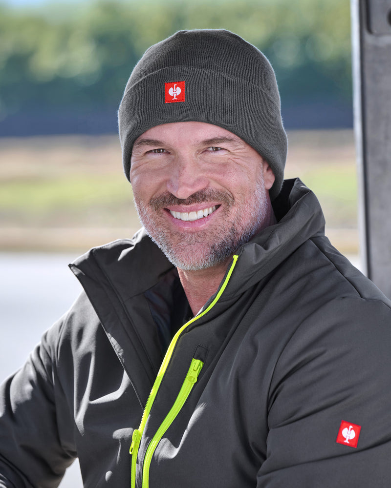 A smiling man wearing a dark gray knit beanie with a red logo patch and a matching dark gray jacket with bright neon green zipper accents, photographed outdoors with a blurred natural background.