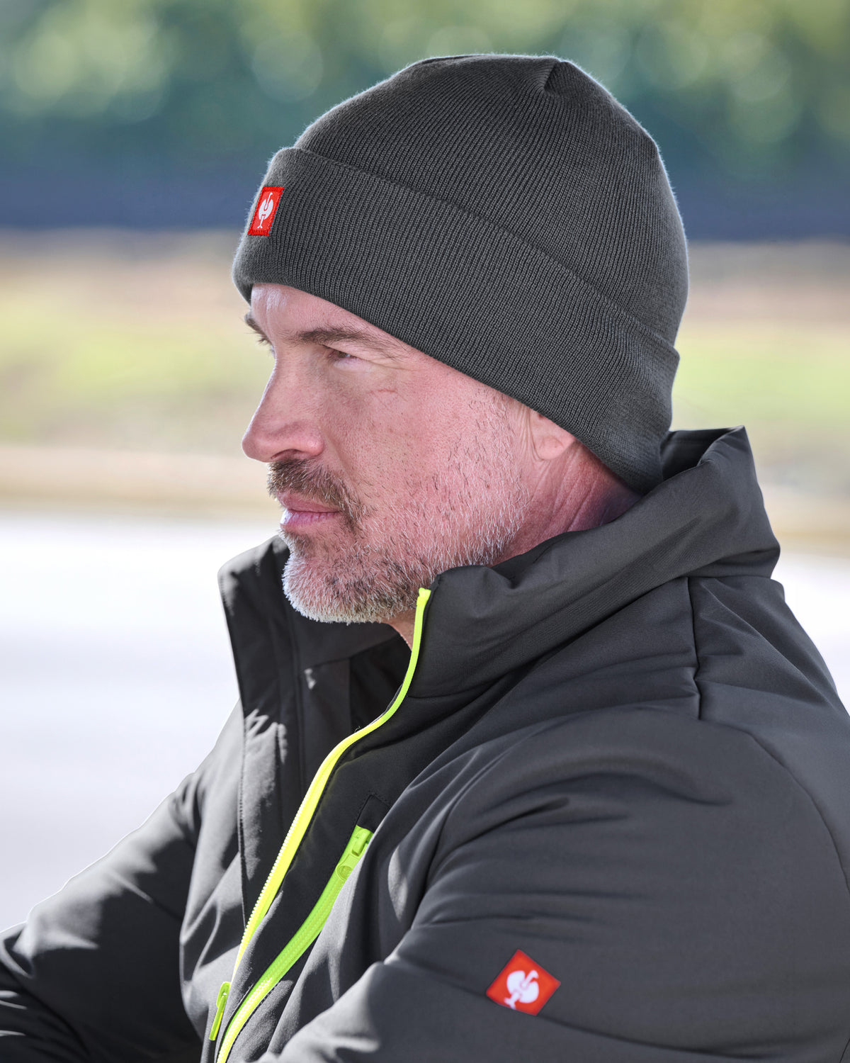 A man in profile wearing a dark gray knit beanie with a small red square logo patch and a black jacket with a bright yellow-green zipper, photographed outdoors with a blurred natural background.