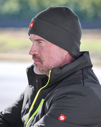A man in profile wearing a dark gray knit beanie with a small red square logo patch and a black jacket with a bright yellow-green zipper, photographed outdoors with a blurred natural background.