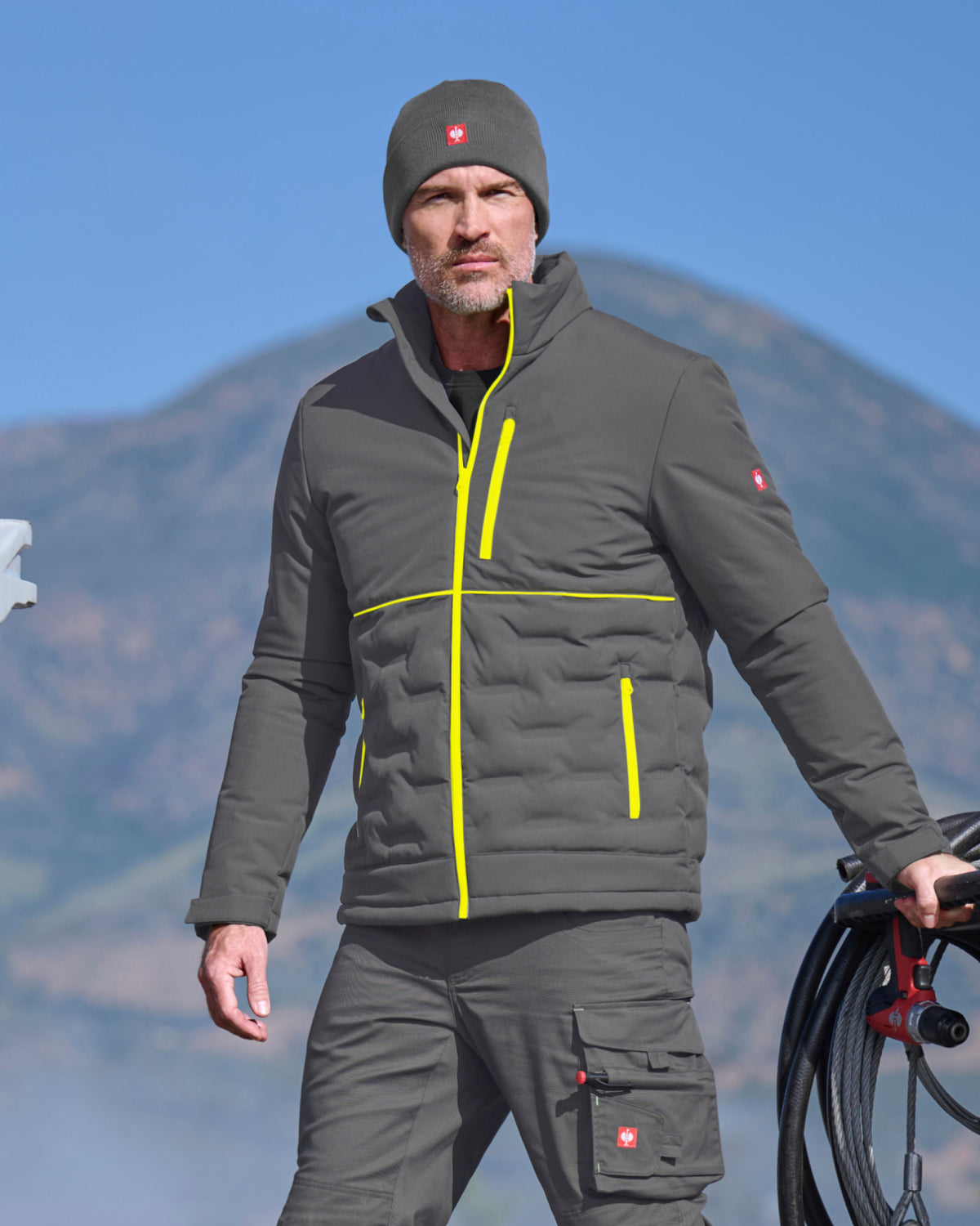 A man wearing a dark gray outdoor jacket with bright yellow zipper accents, matching gray pants, and a gray beanie with a red Swiss cross logo, standing outdoors with a mountain in the background.