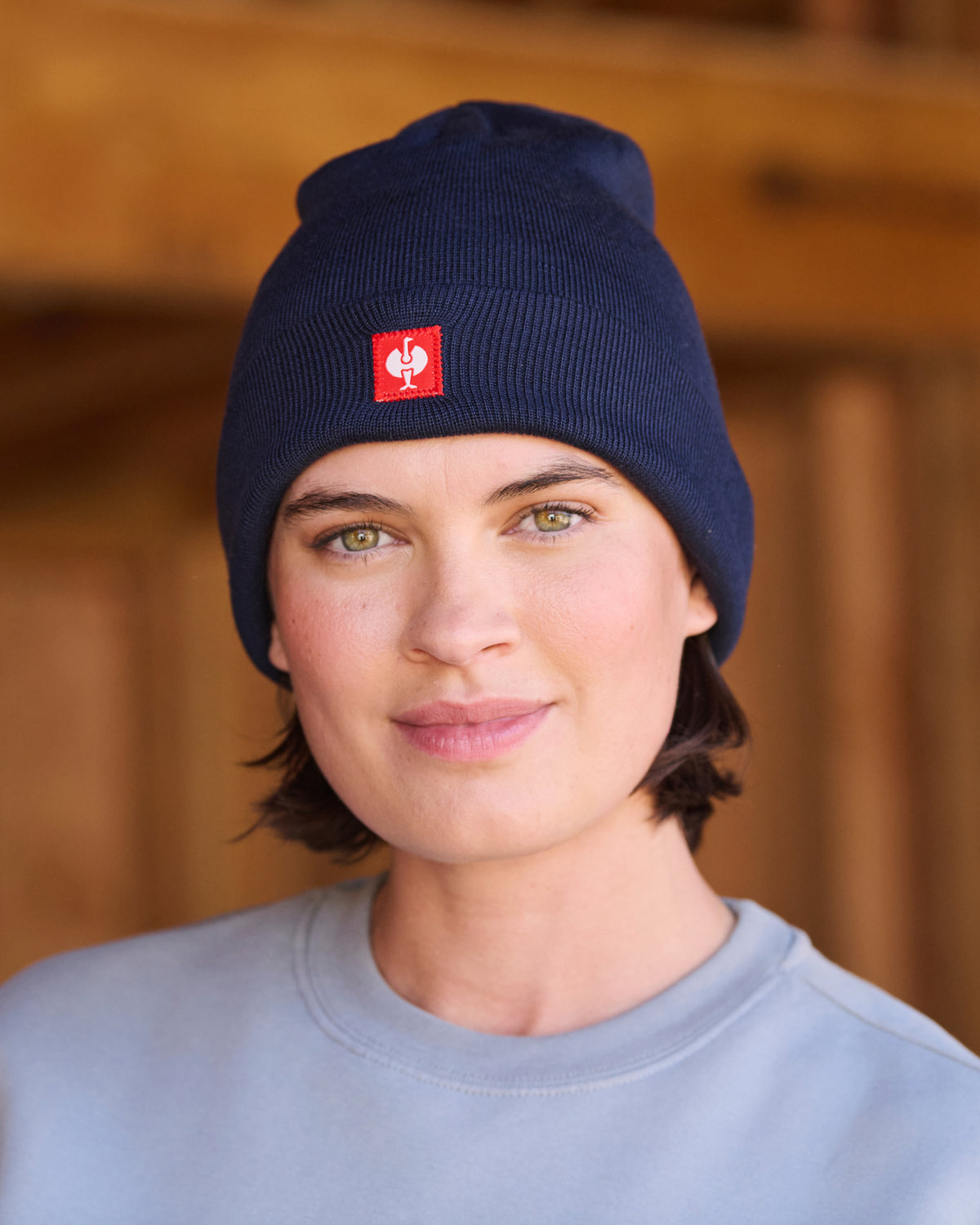 A person wearing a navy blue ribbed knit beanie with a small red square logo patch on the front, photographed against a warm blurred wooden background.