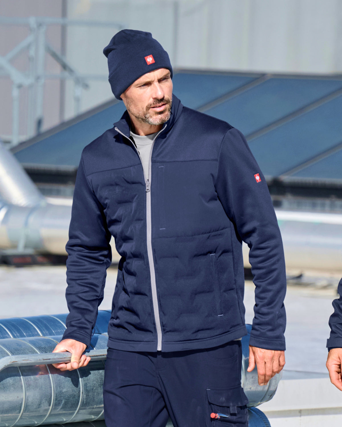 A man wearing a navy blue zip-up jacket, matching navy pants, and a navy beanie with a small red logo, standing outdoors on a rooftop with metal structures and solar panels in the background.
