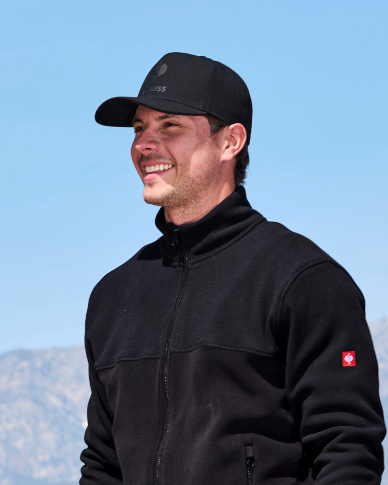 A man wearing a black fleece jacket and a black cap with a small white logo, photographed outdoors against a clear blue sky and blurred mountains.
