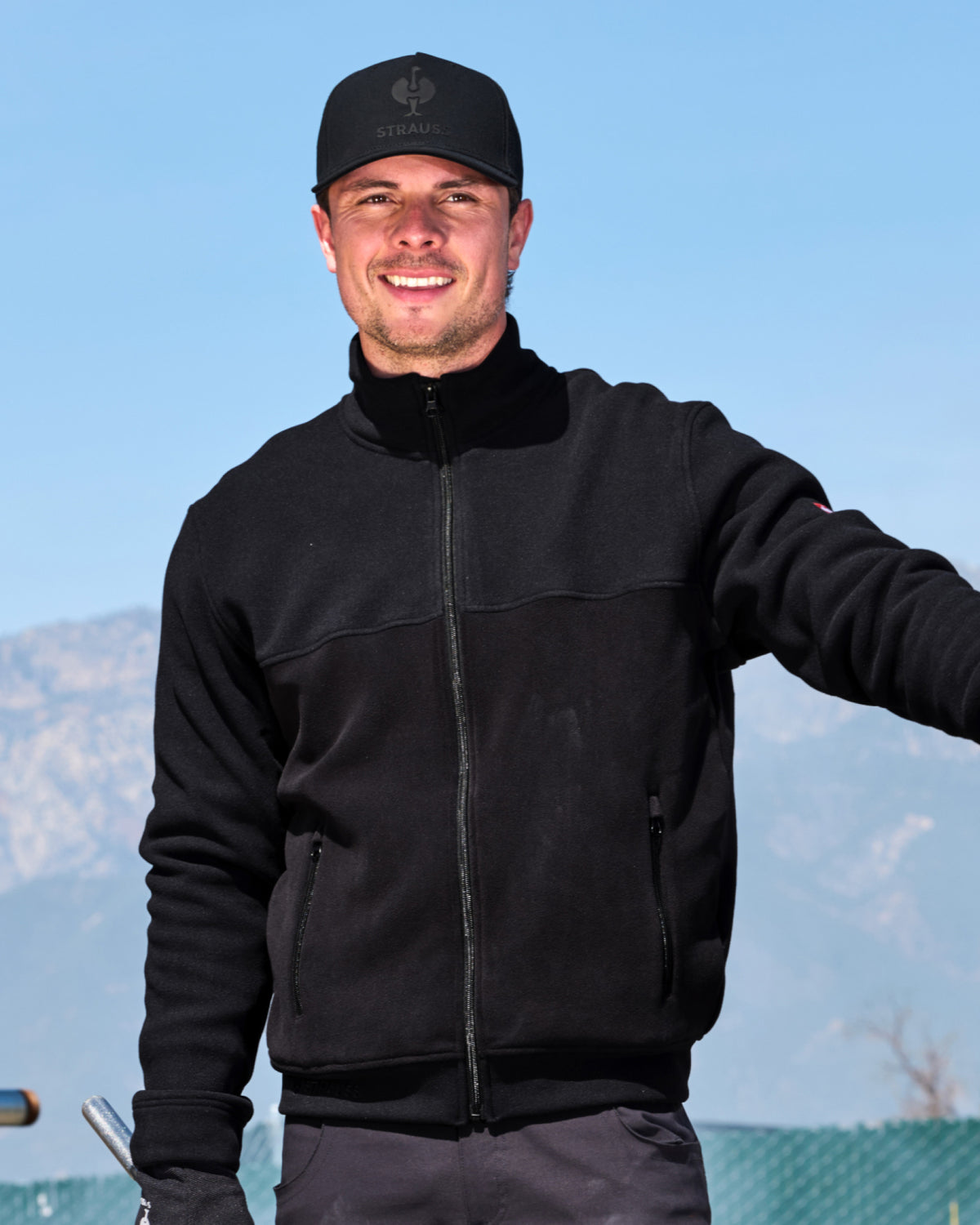 A smiling man wearing a black zip-up fleece jacket, black baseball cap with a visible ZONN logo, and dark pants, standing outdoors against a blue sky and distant mountains.