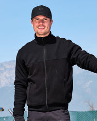 A smiling man wearing a black zip-up fleece jacket, black baseball cap with a visible ZONN logo, and dark pants, standing outdoors against a blue sky and distant mountains.