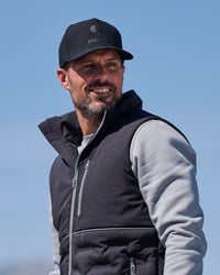 A man wearing a black Stravitz cap, a black zippered puffer vest over a light gray long-sleeve shirt, photographed outdoors against a clear blue sky.