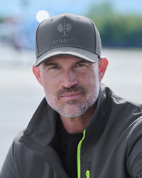 A close-up portrait of a man wearing a dark gray baseball cap with the brand name 'Strauss' and a logo on the front, paired with a dark gray zip-up jacket with a bright neon green zipper.